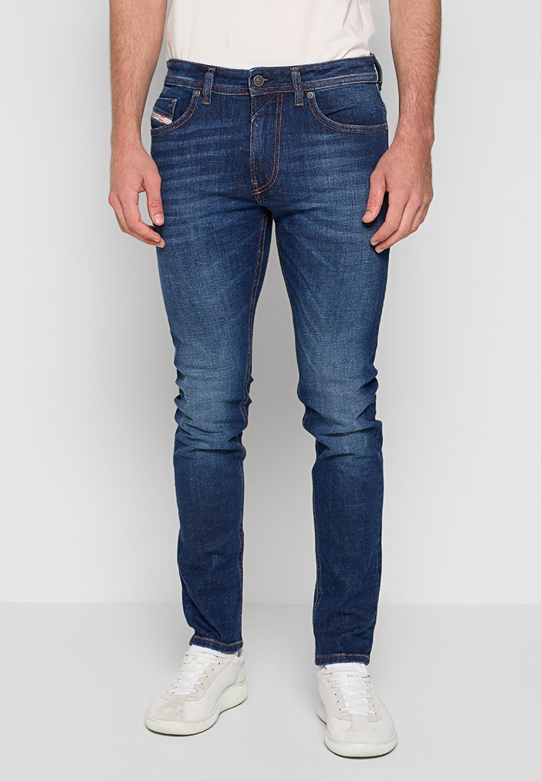 Mann in schmal geschnittenen dunkelblauen Jeans mit dezenten Auswaschungen und weißen Sneakers, vor einem einfarbigen hellen Hintergrund stehend.