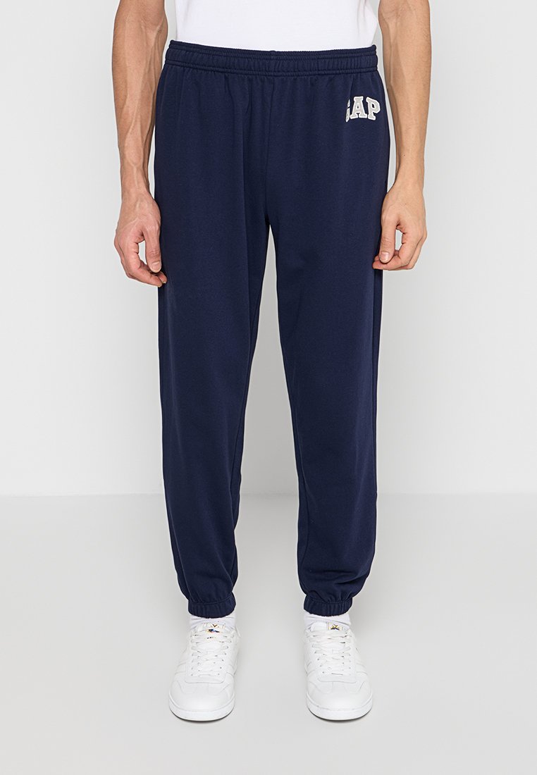 Mann trägt marineblaue Jogginghosen mit elastischem Bund und GAP-Logo am linken Oberschenkel sowie weiße Turnschuhe, steht vor einfachem Hintergrund.
