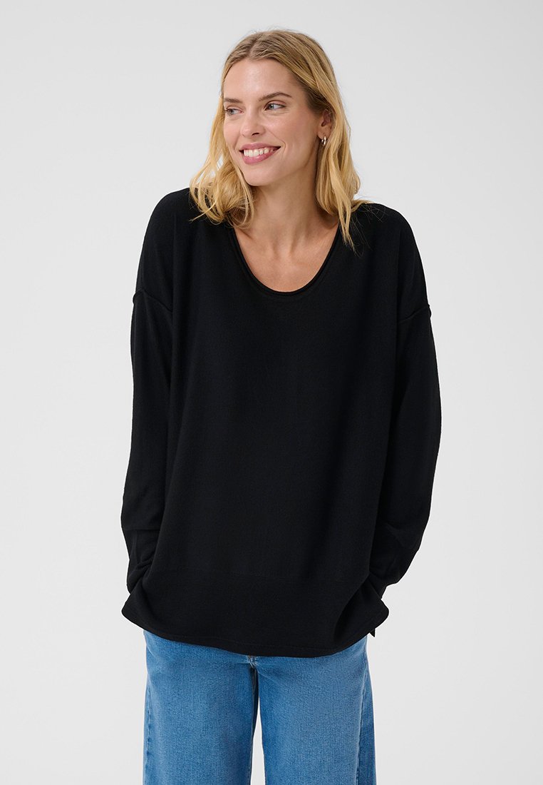 Lächelnde Frau mit blondem Haar trägt ein locker sitzendes schwarzes Langarmshirt und blaue Jeans, die Hände in den Taschen, schaut zur Seite.