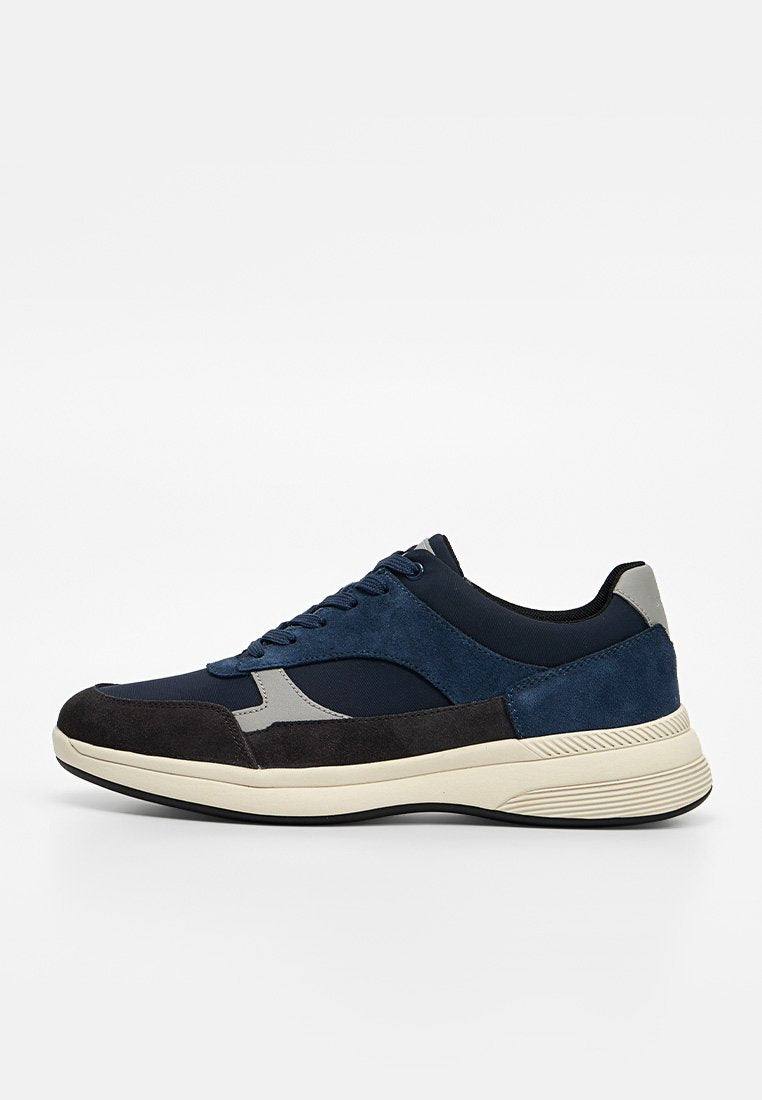 Niedriger Sneaker mit marineblauen, schwarzen und grauen Einsätzen, Schnürverschluss und cremefarbener dicker Sohle auf weißem Hintergrund.