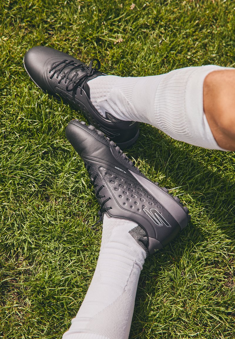 Schwarze Fußballschuhe mit strukturiertem Design, Schnürsenkeln und Gummistollen, kombiniert mit weißen Socken, die auf grünem Gras liegen.