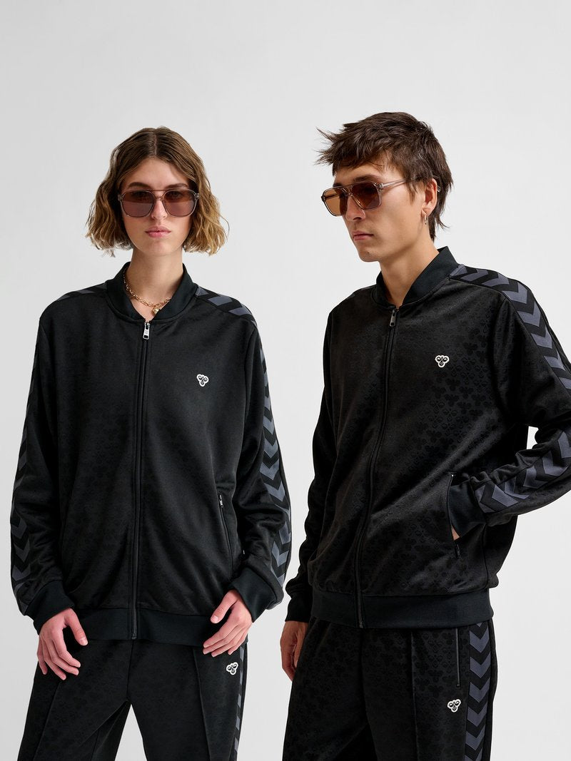Zwei Personen tragen identische schwarze Zip-Up-Trainingsanzüge mit Chevron-Streifen an den Ärmeln und kleinen weißen Logos auf Brust und Hose.