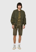 Olivgrüne Bomberjacke mit gesteppter Textur, kombiniert mit einem olivfarbenen Hemd und Shorts. Weiße High-Top-Sneaker vervollständigen das Outfit.