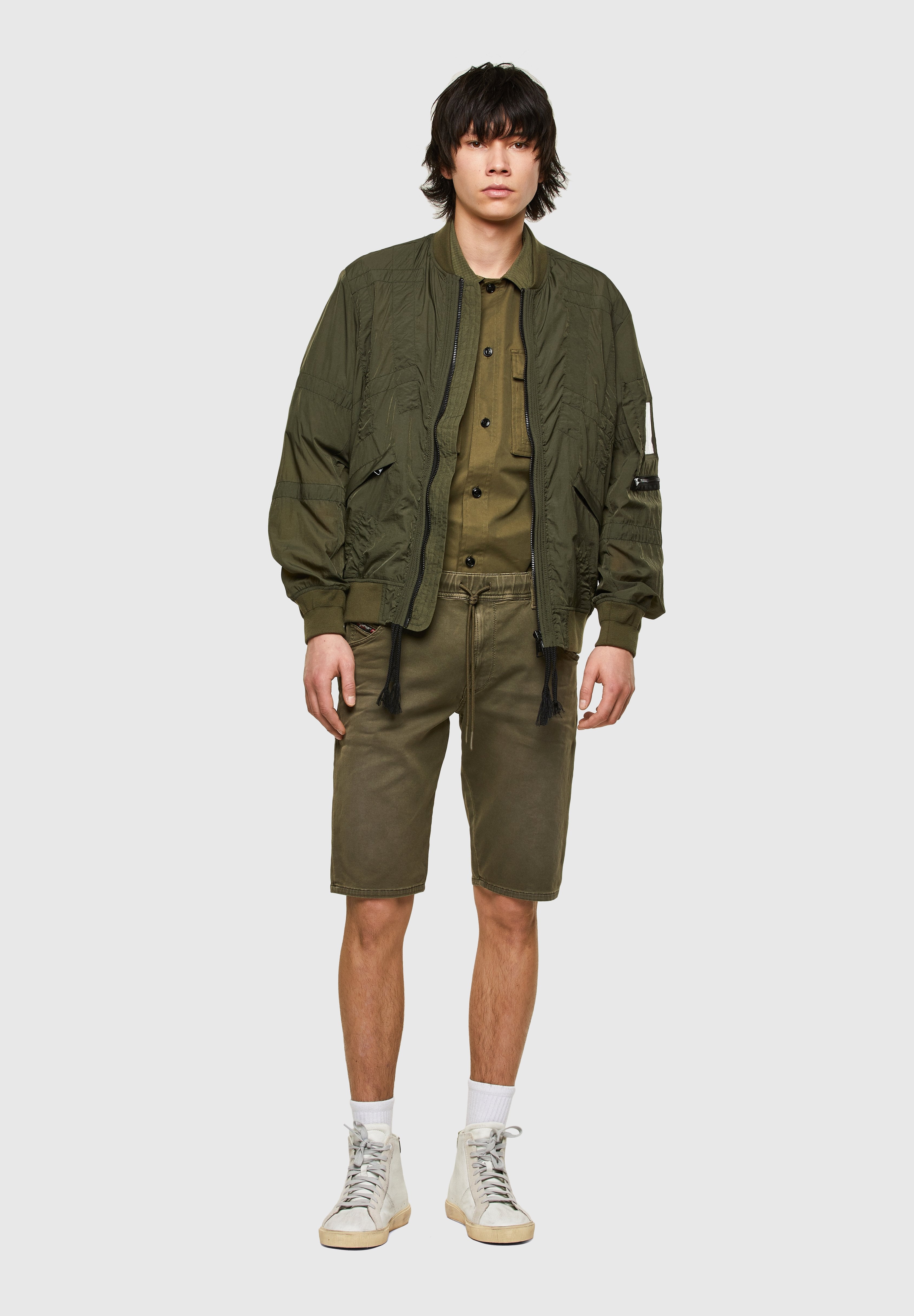 Olivgrüne Bomberjacke mit gesteppter Textur, kombiniert mit einem olivfarbenen Hemd und Shorts. Weiße High-Top-Sneaker vervollständigen das Outfit.