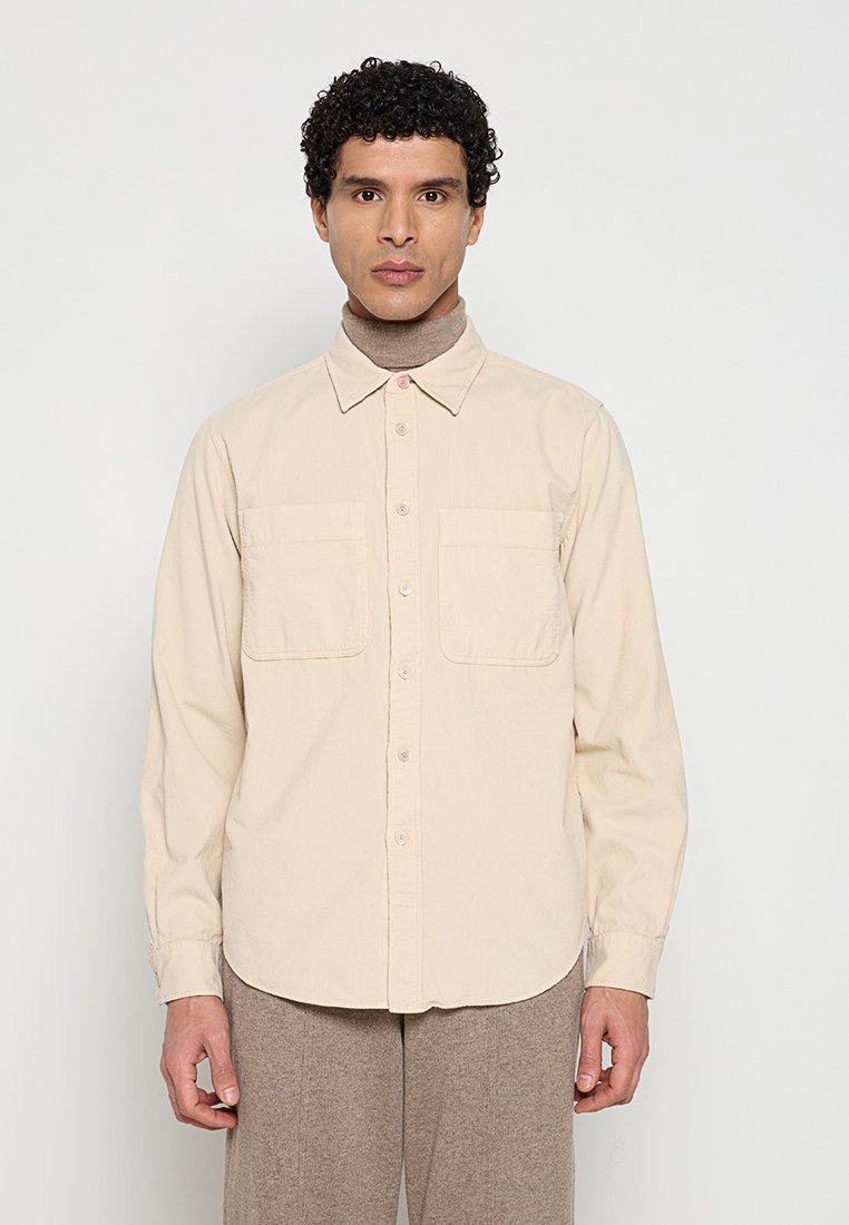 Mann mit lockigem Haar, der ein beigefarbenes Hemd über einem braunen Rollkragenpullover und braune Hosen trägt, vor einem schlichten weißen Hintergrund steht.