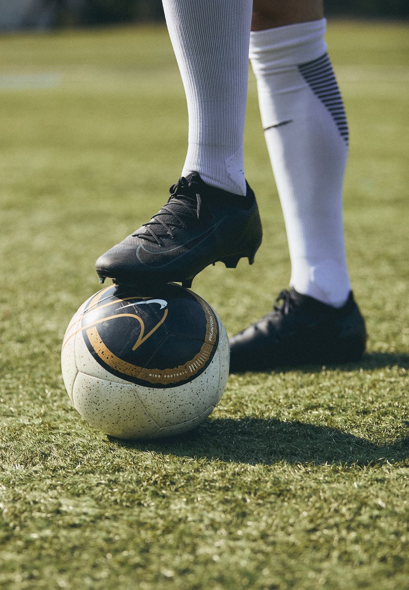 Schwarze Fußballschuhe auf einem weiß-schwarz gemusterten Fußball, der auf grünem Gras ruht. Der Schuh hat eine strukturierte Oberfläche und Schnürsenkel.