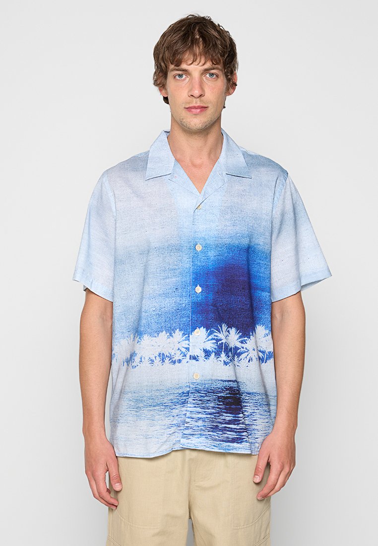 Junger Mann trägt ein kurzärmeliges Hemd mit Knöpfen und blau-weißem tropischem Strand- und Palmenmotiv, steht vor einem einfarbigen Hintergrund.