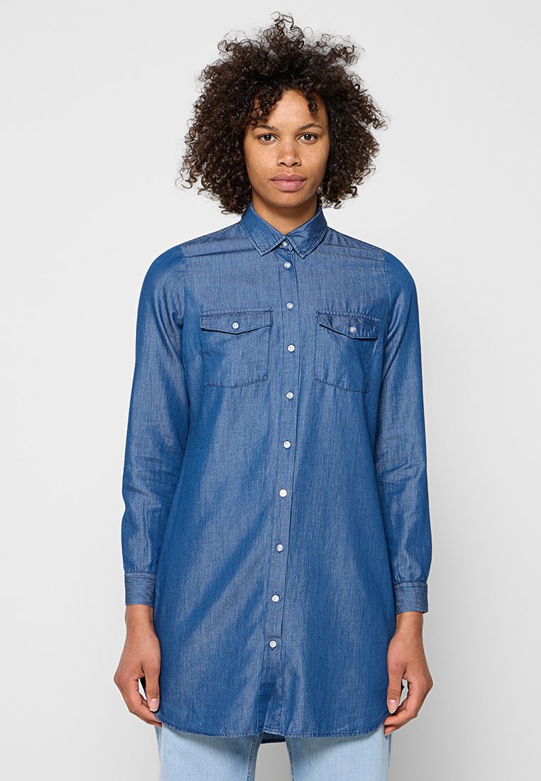 Person mit lockigem Haar, die ein langärmeliges blaues Jeanshemd mit Brusttaschen trägt, steht vor einem einfarbigen hellen Hintergrund.