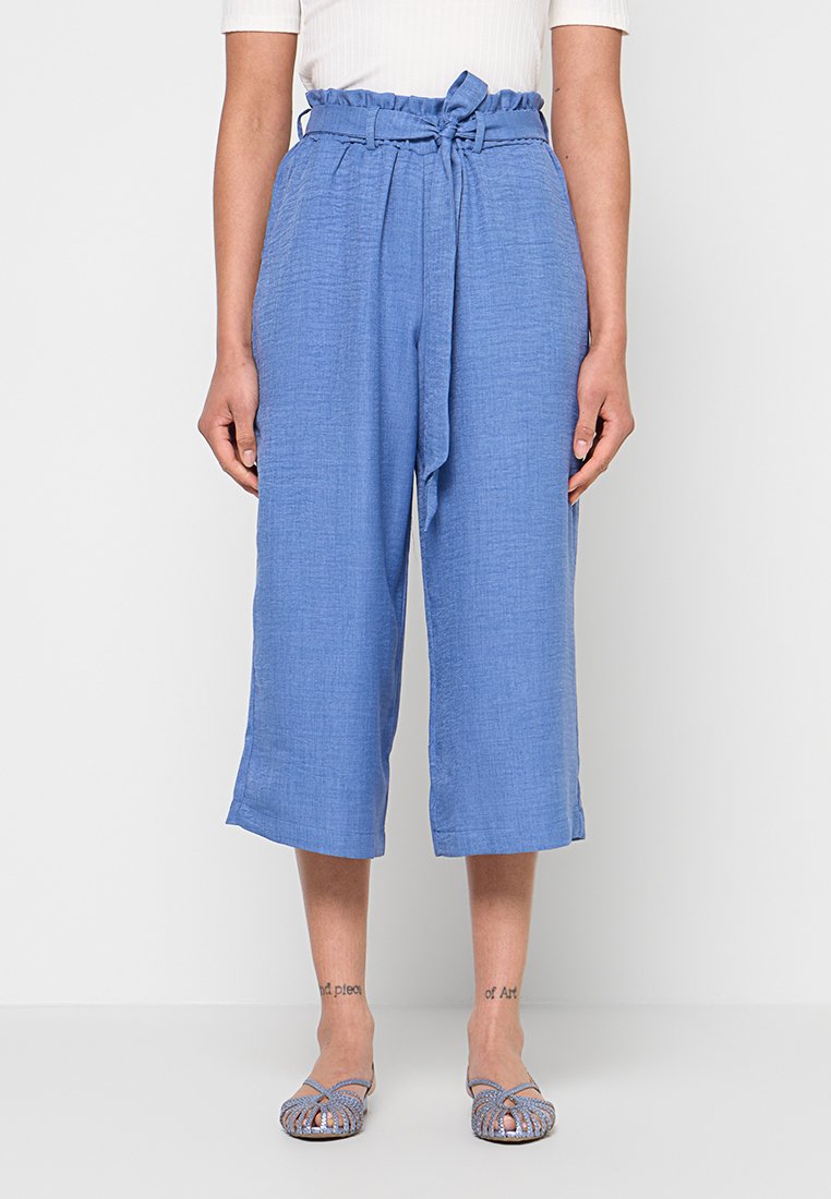 Frau trägt blaue, hoch taillierte, cropped Hosen mit einem gebundenen Gürtel, kombiniert mit blauen, gewebten flachen Schuhen, stehend vor weißem Hintergrund.