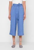 Frau trägt blaue, hoch taillierte, cropped Hosen mit einem gebundenen Gürtel, kombiniert mit blauen, gewebten flachen Schuhen, stehend vor weißem Hintergrund.