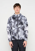 Junger Mann trägt eine schwarz-weißes Batik-Reißverschlussjacke mit Kapuze und schwarze Hose, steht vor einem schlichten hellen Hintergrund.