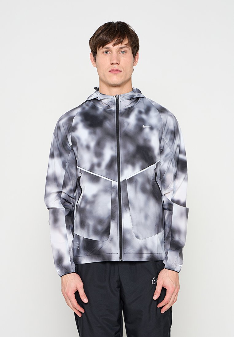 Junger Mann trägt eine schwarz-weißes Batik-Reißverschlussjacke mit Kapuze und schwarze Hose, steht vor einem schlichten hellen Hintergrund.