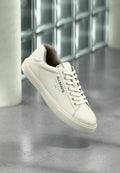 Weißer AllSaints-Sneaker mit Schnürsenkeln, Logo und japanischem Text, der über einem polierten Betonboden mit mattierten Glasbausteinen im Hintergrund schwebt.