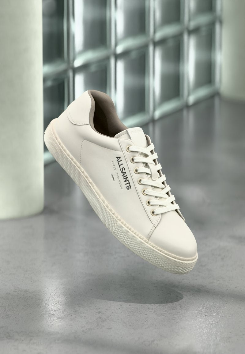 Weißer AllSaints-Sneaker mit Schnürsenkeln, Logo und japanischem Text, der über einem polierten Betonboden mit mattierten Glasbausteinen im Hintergrund schwebt.
