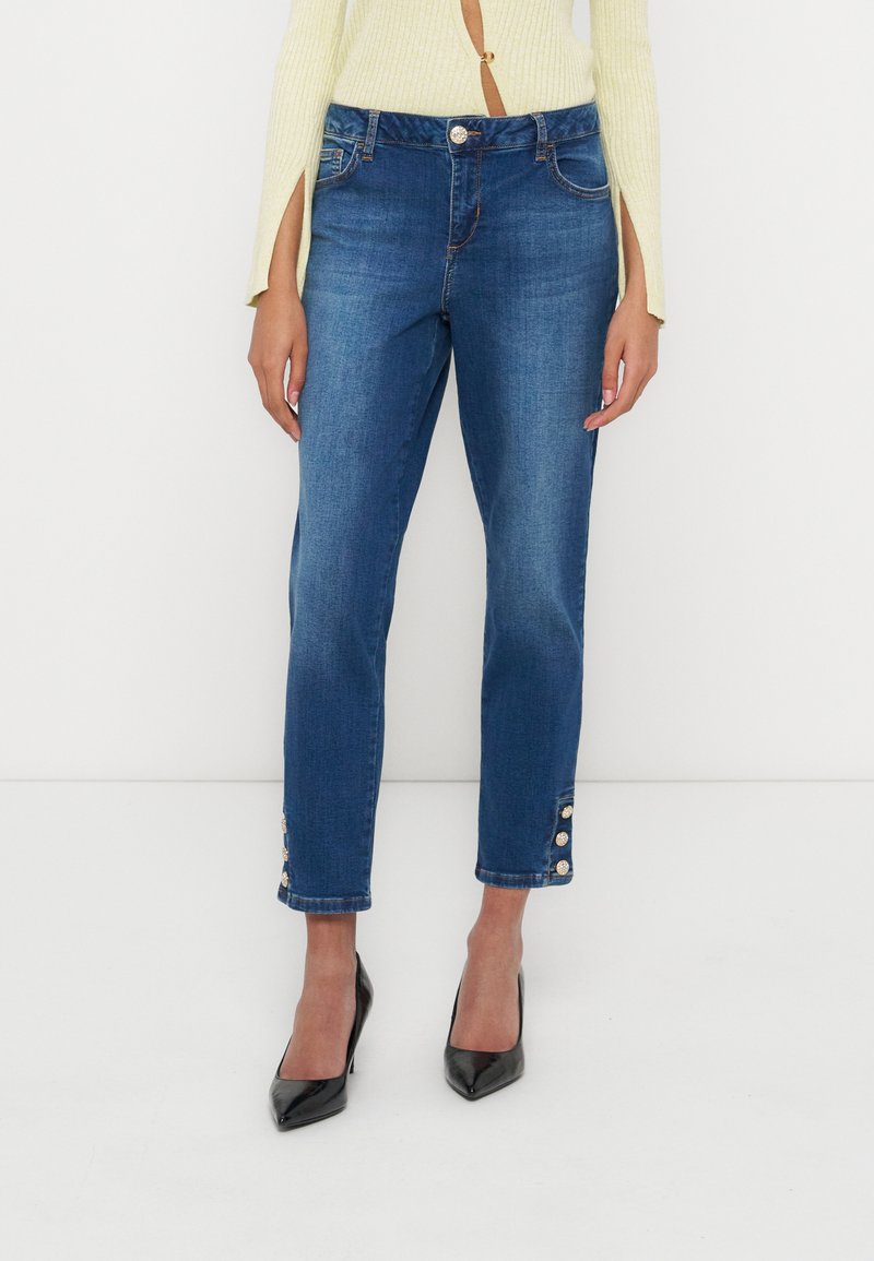 Frau trägt blaue Jeans mit geradem Bein und Knopfdetails am Knöchel, einen cremefarbenen gerippten Pullover und schwarze spitze High Heels.