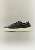 Schwarzer Sneaker mit niedrigem Schnitt, schwarzen Schnürsenkeln, weißer Sohle und goldfarbenem "HUGO"-Logo an der Seite und Ferse vor einfachem Hintergrund.