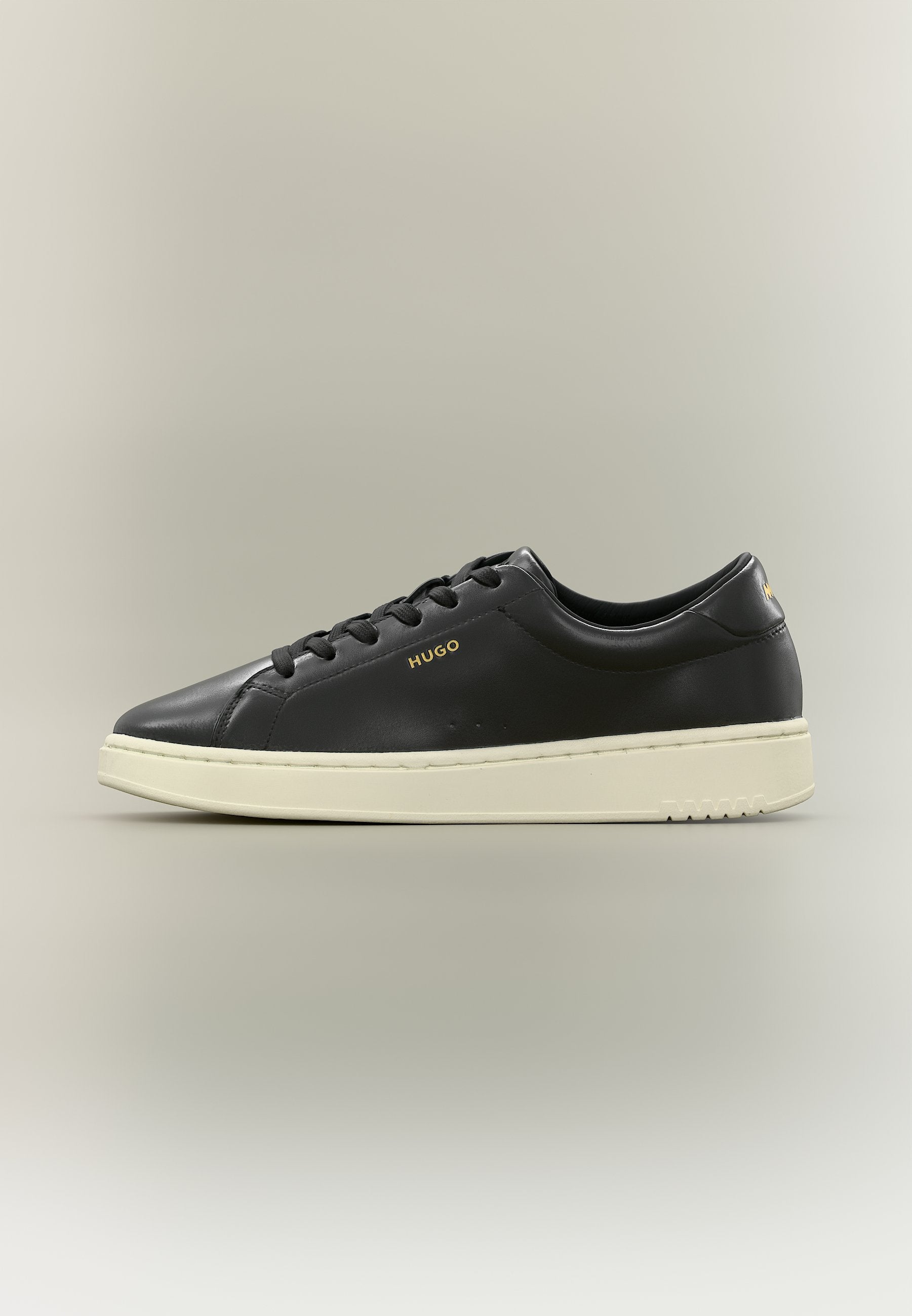 Schwarzer Sneaker mit niedrigem Schnitt, schwarzen Schnürsenkeln, weißer Sohle und goldfarbenem "HUGO"-Logo an der Seite und Ferse vor einfachem Hintergrund.