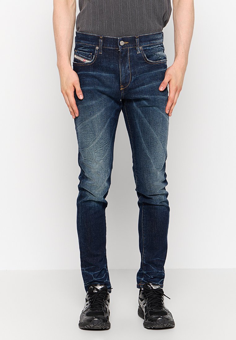 Mann in dunkelblauen Slim-Fit-Jeans mit ausgebleichten Details und schwarzen Sneakern, steht vor einem schlichten weißen Hintergrund.