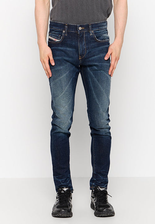 Mann in dunkelblauen Slim-Fit-Jeans mit ausgebleichten Details und schwarzen Sneakern, steht vor einem schlichten weißen Hintergrund.