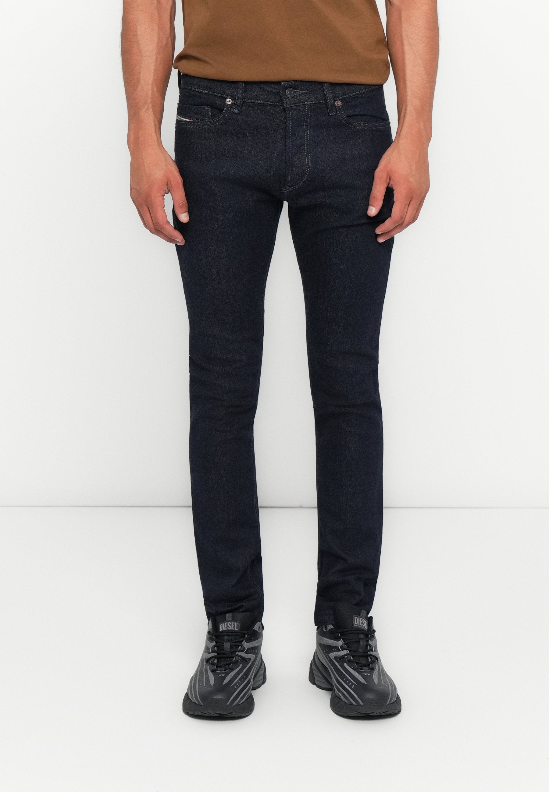 Mann in dunkelblauen Slim-Fit-Jeans und schwarzen Diesel-Sneakern, der vor einem schlichten weißen Hintergrund steht.