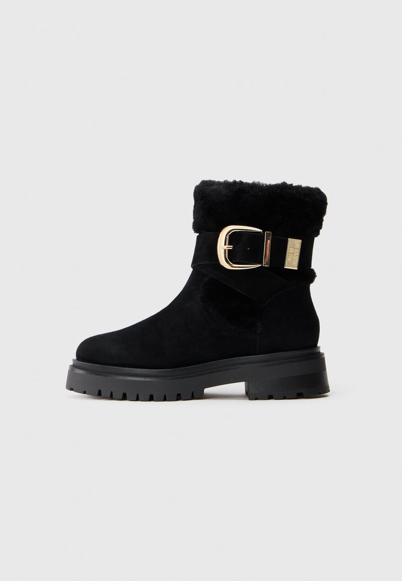 Schwarze Wildleder-Stiefelette mit dicker Gummisohle, gefaltetem Lammfellsaum und großem goldenen Schnallenriemendetail.