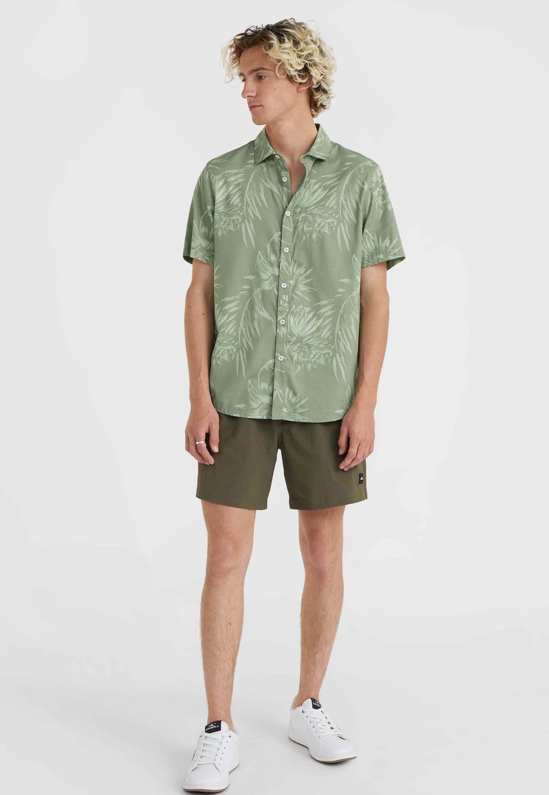 Kurzärmliges, hellgrünes Hemd mit Knopfleiste und tropischem Blattmuster, kombiniert mit olivgrünen Shorts und weißen Sneakers.