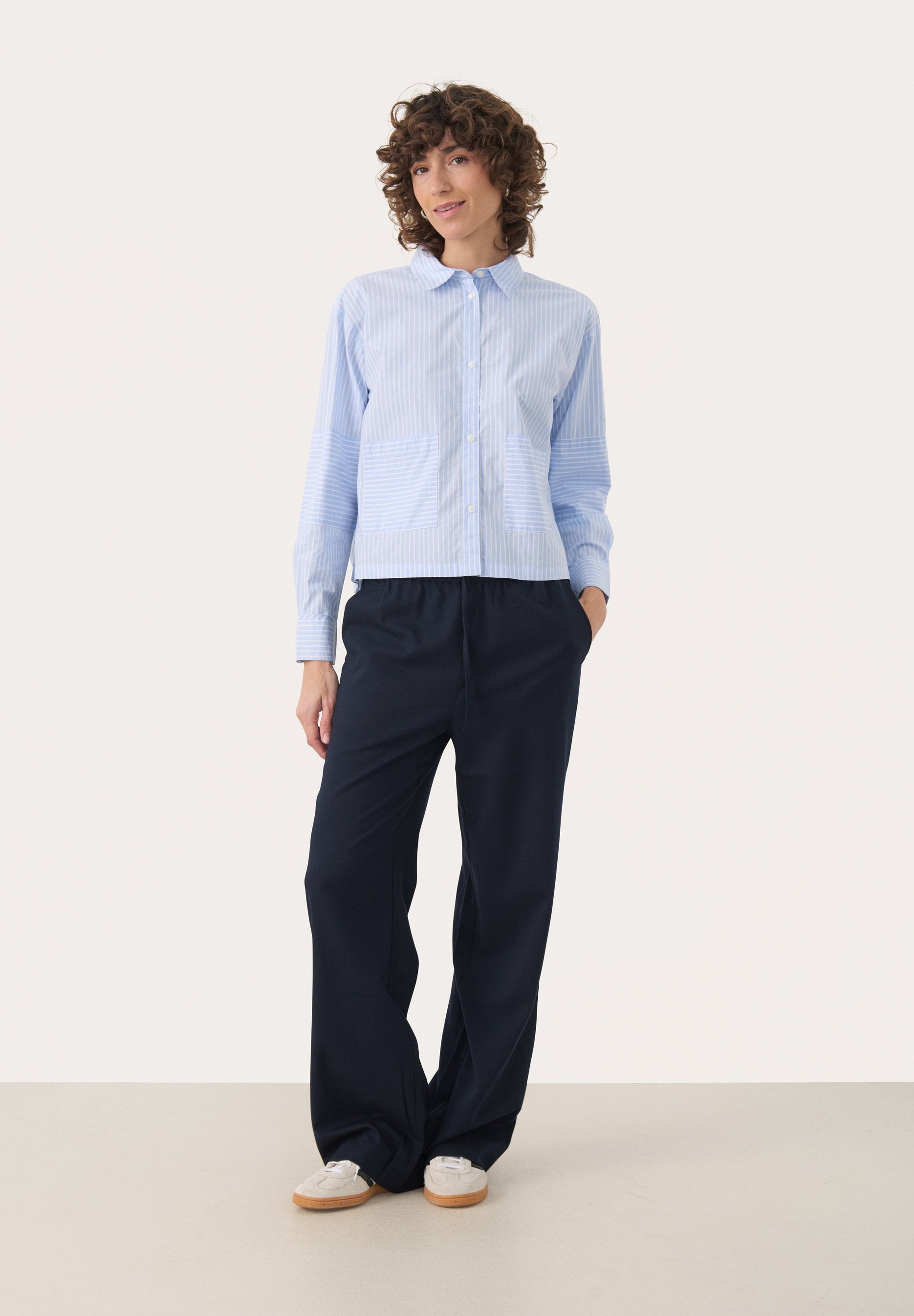 Hellblaues Button-up-Hemd mit vertikalen Streifen und zwei Fronttaschen, kombiniert mit dunkelblauen weiten Hosen und weißen Sneakers.