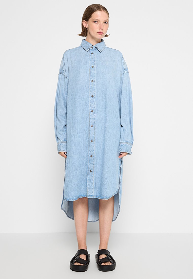 Frau, die ein hellblaues Langarm-Denim-Kleiderhemd mit vorderem Knopfverschluss und schwarzen Plateau-Sandalen trägt, steht vor einer schlichten Wand.