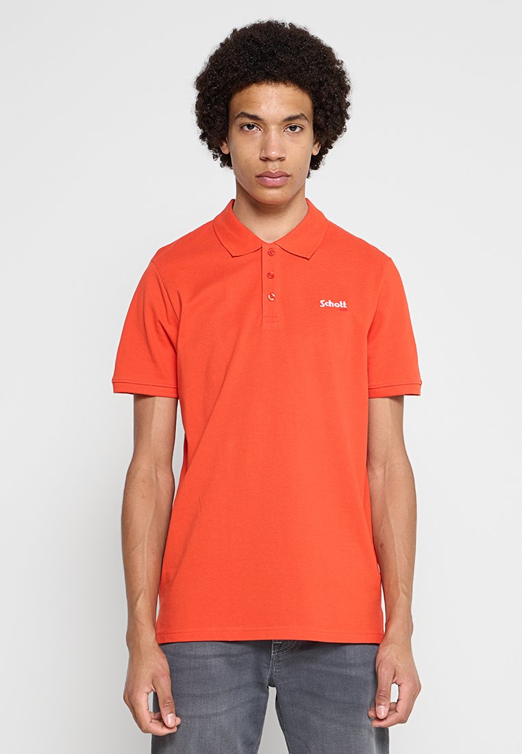 Junger Mann mit lockigem Haar, der ein oranges Poloshirt und graue Jeans trägt, steht vor einem schlichten hellgrauen Hintergrund.