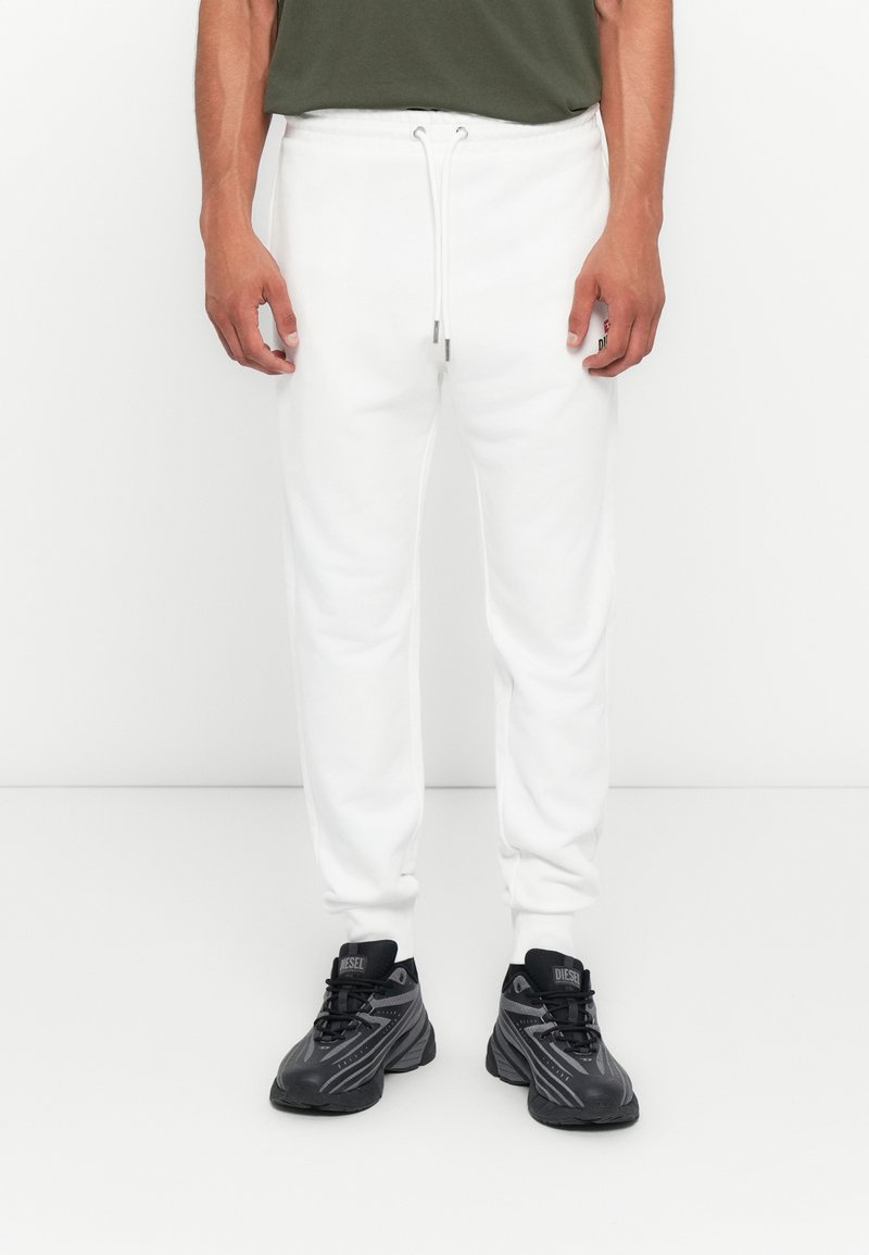 Mann in weißen Jogginghosen mit elastischem Bund und Kordelzug, dunklen Sneakers und einem olivgrünen Shirt, steht vor einem weißen Hintergrund.
