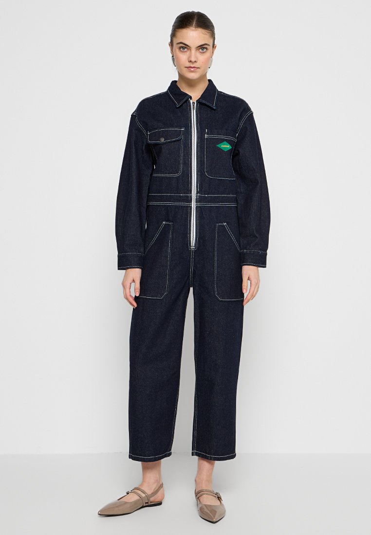 Denim-Overall mit einem Reißverschluss vorne, langen Ärmeln, zwei Brusttaschen und kontrastierenden weißen Nähten. Dunkelblaue Farbe, entspannter Schnitt.