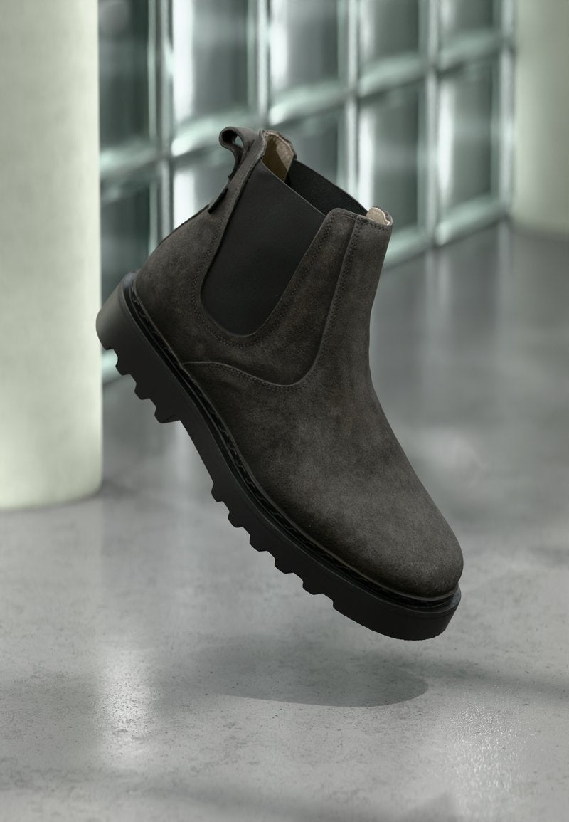 Dunkelgrauer Chelsea-Stiefel aus Wildleder mit schwarzen elastischen Seitenteilen und dicker schwarzer Profilsohle auf reflektierendem Betonboden.