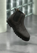 Dunkelgrauer Chelsea-Stiefel aus Wildleder mit schwarzen elastischen Seitenteilen und dicker schwarzer Profilsohle auf reflektierendem Betonboden.