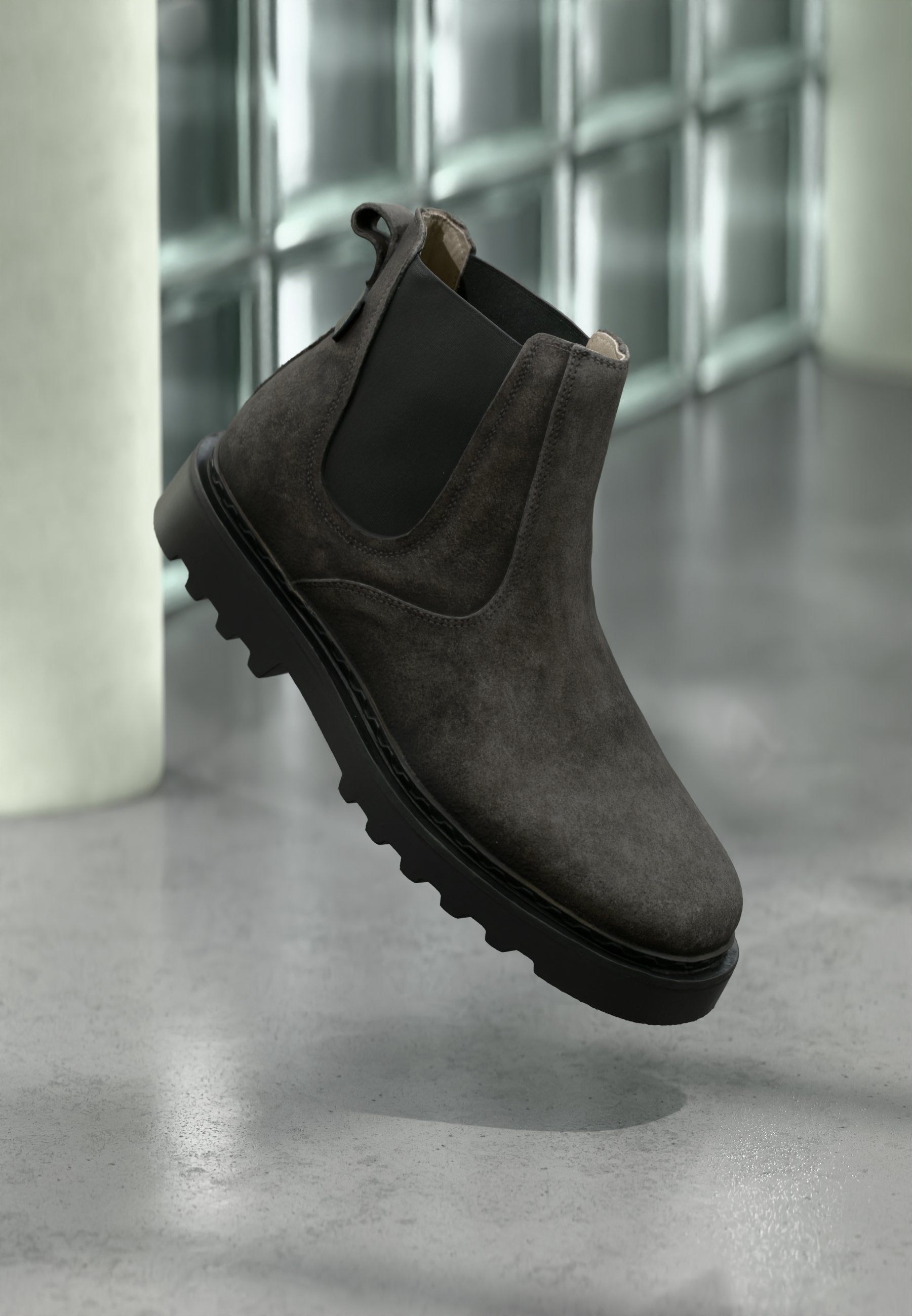 Dunkelgrauer Chelsea-Stiefel aus Wildleder mit schwarzen elastischen Seitenteilen und dicker schwarzer Profilsohle auf reflektierendem Betonboden.