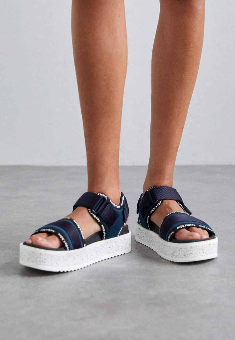 Marineblaue Sandalen mit Velcro-Riemen und weiß gesprenkelten Sohlen. Offenes Toe-Design mit kontrastierenden dunklen Details und Markenakzenten.