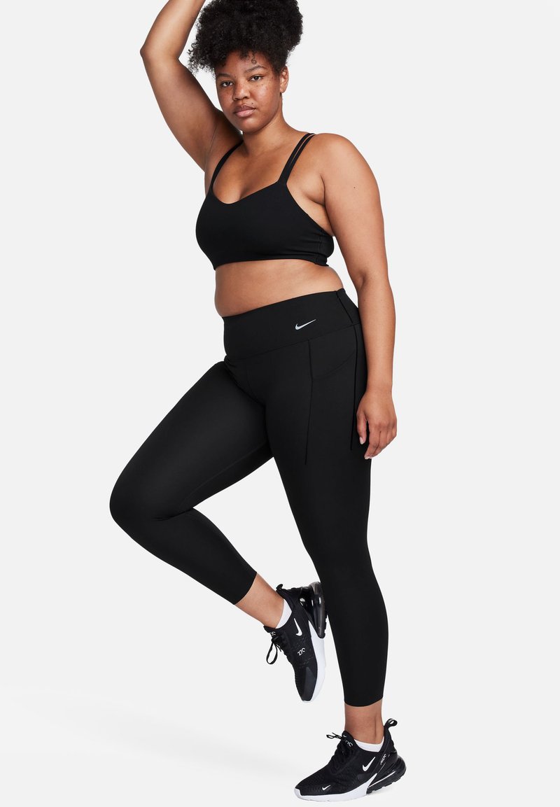 Schwarz Sportbekleidungsset mit einem gefütterten, kurzen Sport-BH und hochgeschnittenen Leggings mit einem Logo. Das Modell trägt schwarze Sneakers.