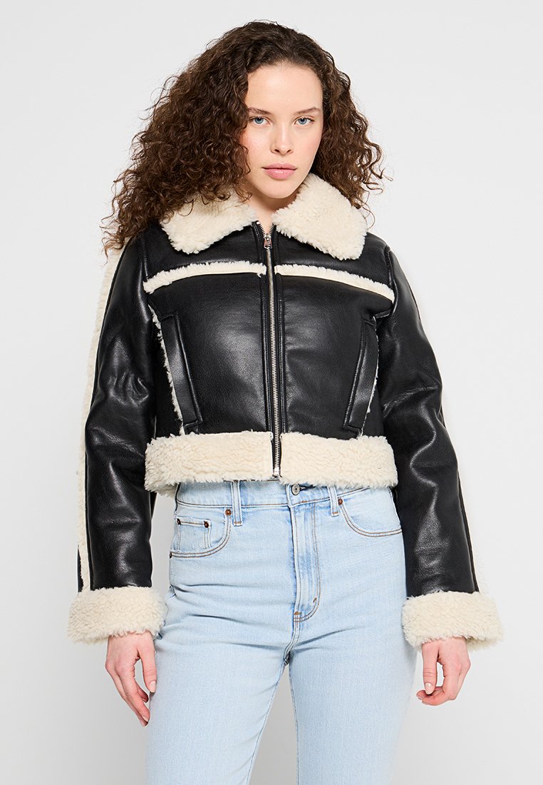 Schwarze Lederjacke mit kurzem Schnitt und Schafslederkragen und -manschetten, ausgestattet mit einem Reißverschluss vorne und breiten horizontalen Streifen. Die Jeans aus Denim vervollständigen das Outfit.