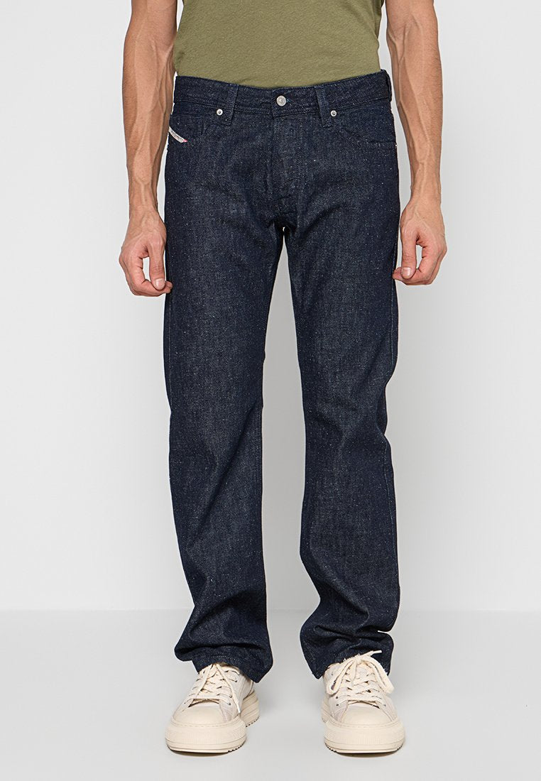 Dunkelblaue Jeans mit geradem Schnitt, ausgestattet mit einem klassischen Fünf-Taschen-Design, Knopfverschluss und dezenten Sprenkeln. Kombiniert mit beigen Sneakers.