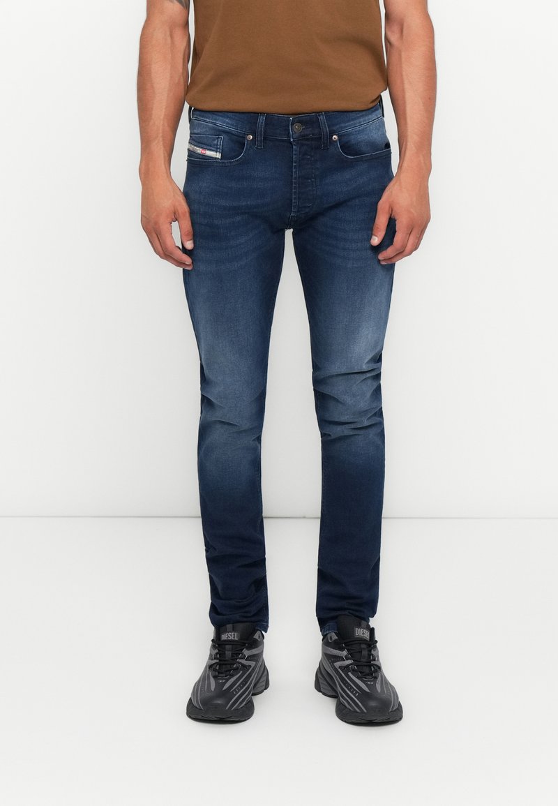 Mann in schmal geschnittenen, dunkelblauen, ausgewaschenen Jeans, braunem Hemd und schwarzen Diesel-Sneakern, steht vor einem weißen Hintergrund.