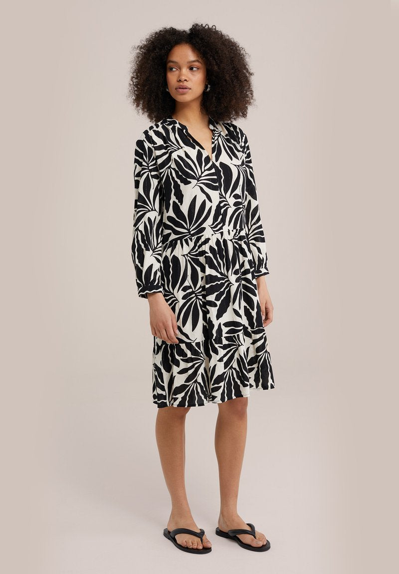 Schwarz-weißes, blumiges Kleid mit langen Ärmeln, V-Ausschnitt, gestuftem Saum und lockerer Passform aus leichtem Stoff. Getragen mit schwarzen Sandalen.