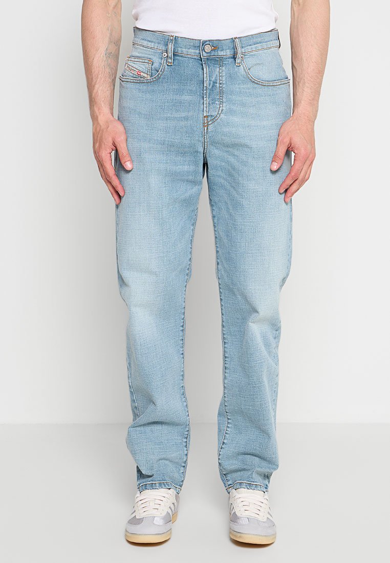 Mann trägt hellblaue Jeans mit geradem Bein und weiße Sneakers, steht mit entspannten Händen seitlich an einem einfarbigen Hintergrund.