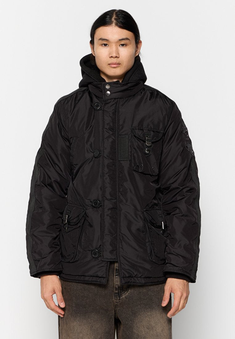 Junge Person mit langen dunklen Haaren trägt eine schwarze, gepolsterte Jacke mit mehreren Vorder- taschen, Knöpfen und einer Reißverschlusskapuze über braunen Hosen.