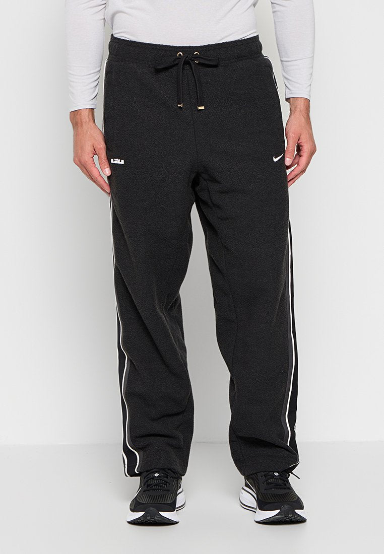 Mann trägt schwarze Nike-Sweatpants mit weißen Seitenstreifen, einem Kordelzug in der Taille und schwarzen und weißen Laufschuhen, steht vor einem weißen Hintergrund.