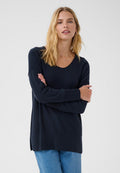 Blonde Frau mit offenem Haar, die einen dunkelblauen Pullover mit langen Ärmeln und hellblaue Jeans trägt, steht mit verschränkten Armen.