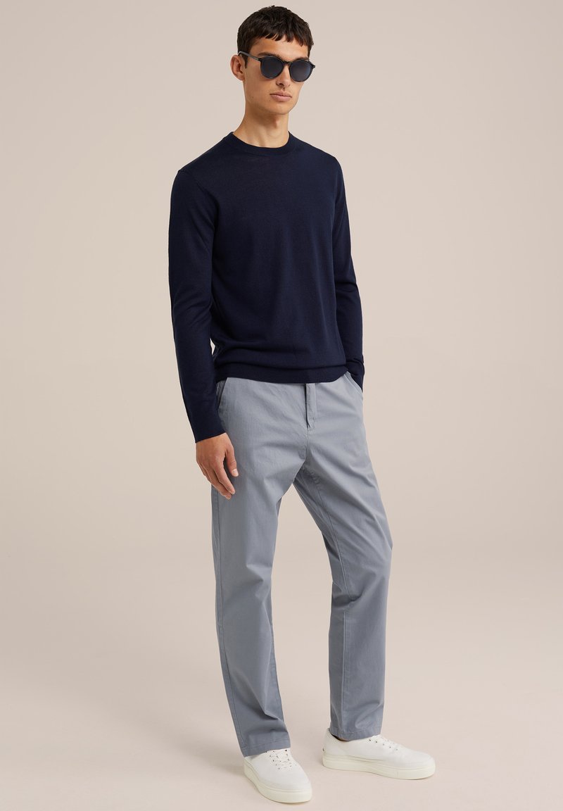 Marineblauer Rundhals-Pullover, hellblaue Chinos und weiße Sneakers. Das Modell trägt runde, schwarze Sonnenbrillen. Glatte Textur, lässige Passform und klare Linien.