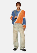 Junge Person trägt einen blau-orange gestreiften Pullover aus Kabelstrick, hellbeige Hosen, schwarze Schuhe und hält einen gestreiften Taschenriemen über eine Schulter.