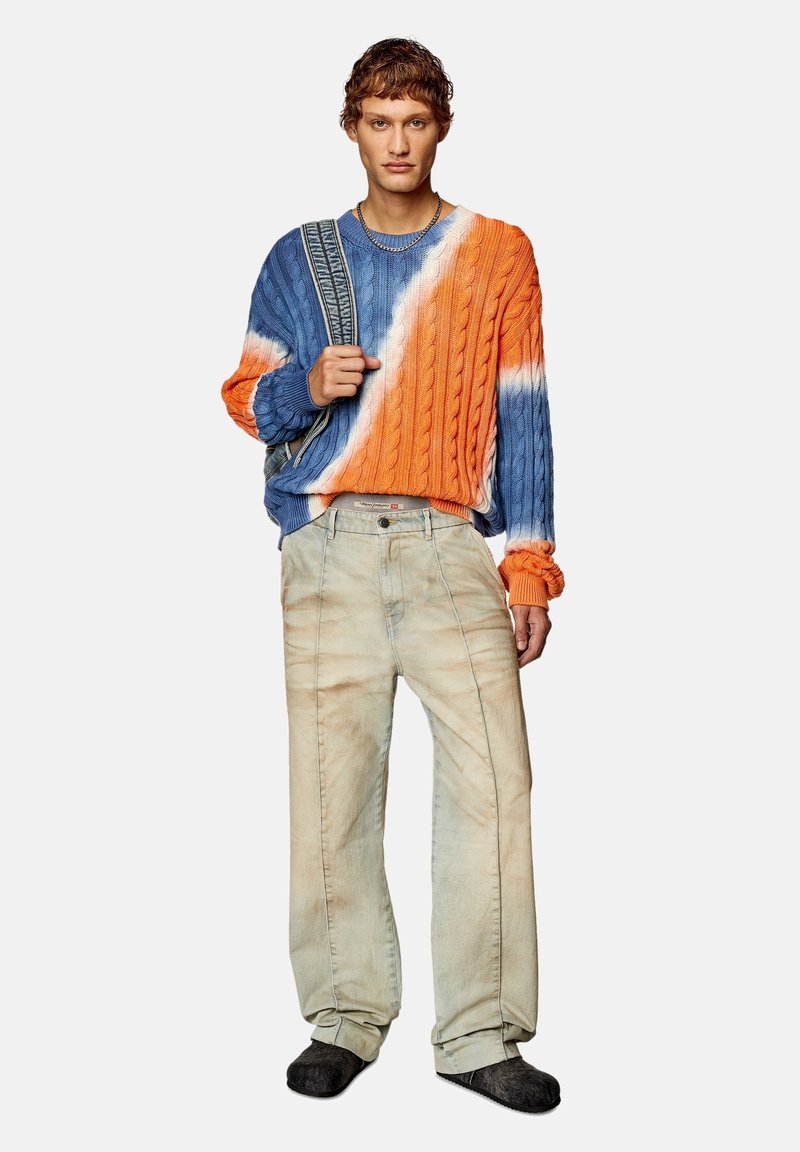 Junge Person trägt einen blau-orange gestreiften Pullover aus Kabelstrick, hellbeige Hosen, schwarze Schuhe und hält einen gestreiften Taschenriemen über eine Schulter.