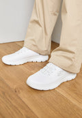Weiße Netz-Sportschuhe mit strukturiertem Obermaterial und elastischen Schnürsenkeln. Die Sohle ist gepolstert und hat ein Wellenmuster. Getragen mit beigen Hosen auf einem Holzboden.