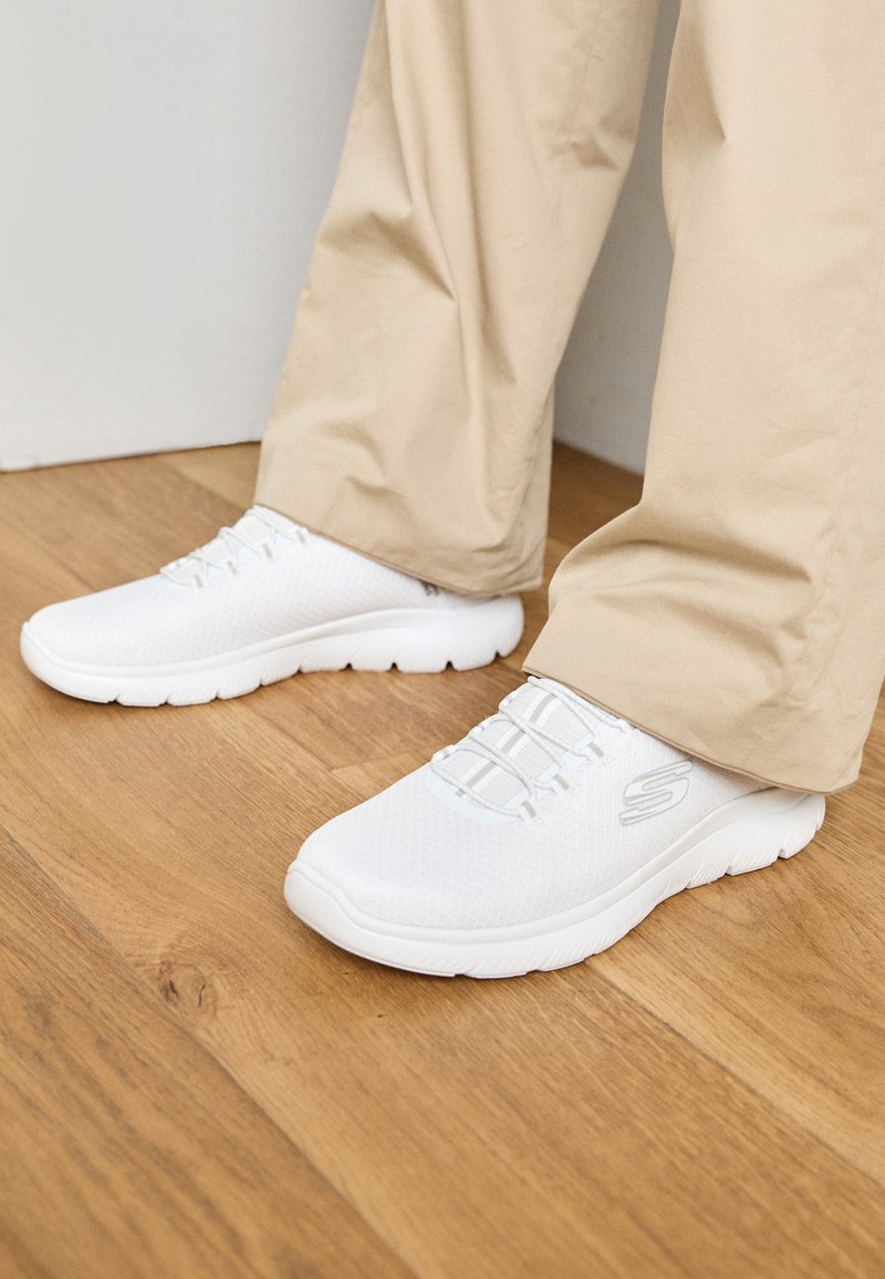Weiße Netz-Sportschuhe mit strukturiertem Obermaterial und elastischen Schnürsenkeln. Die Sohle ist gepolstert und hat ein Wellenmuster. Getragen mit beigen Hosen auf einem Holzboden.