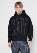 Junger Mann trägt schwarzen Hoodie mit großem aufgenähtem "XXIV"-Motiv auf der Vorderseite und dunkle gemusterte Hose, steht vor schlichtem grauem Hintergrund.